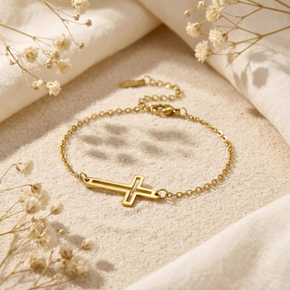 Gold cutout cross chain bracelet shown up close on sand beside soft white fabric and dried floral sprigs.