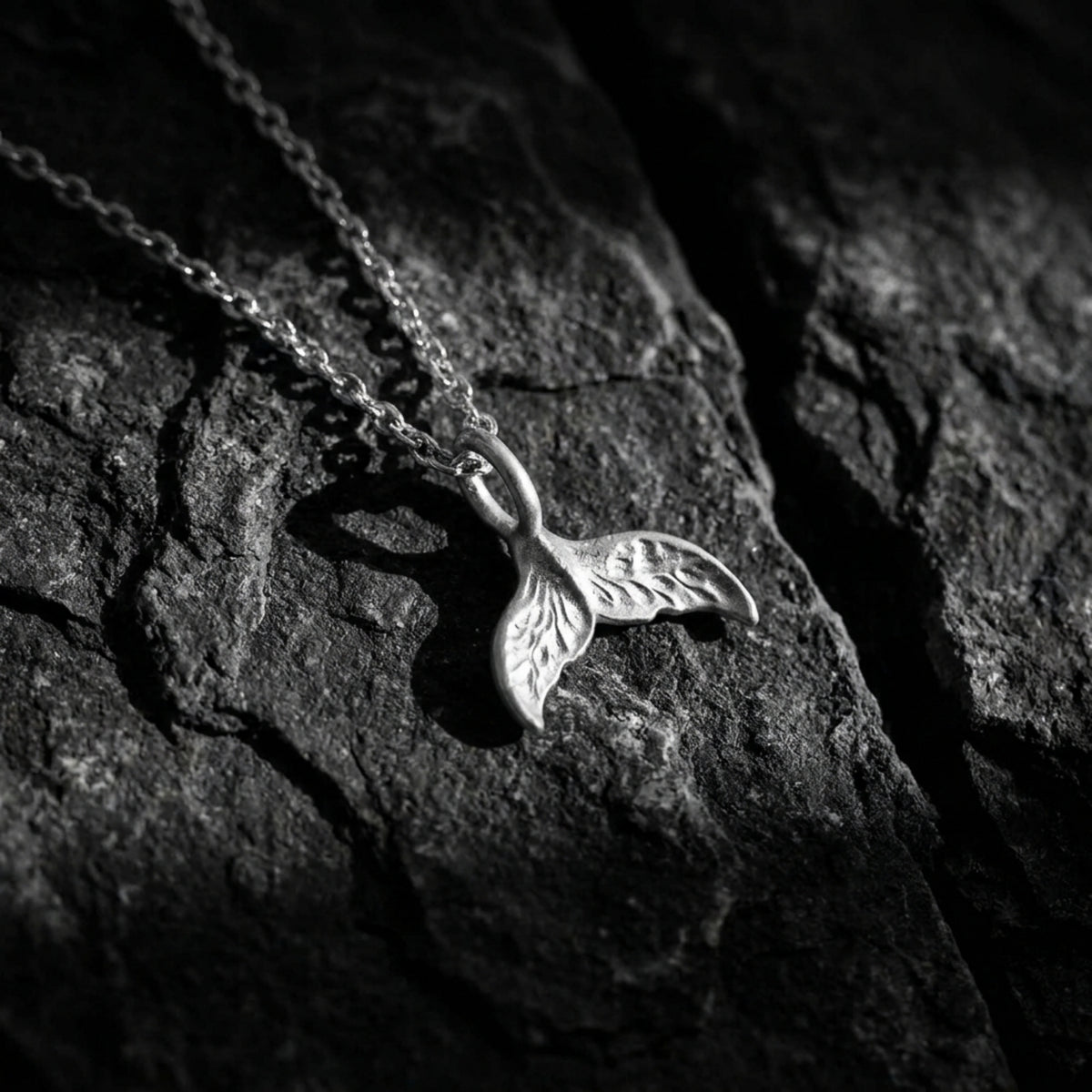 Angled close-up of a silver whale tail pendant and chain resting on a dark, textured rock surface.