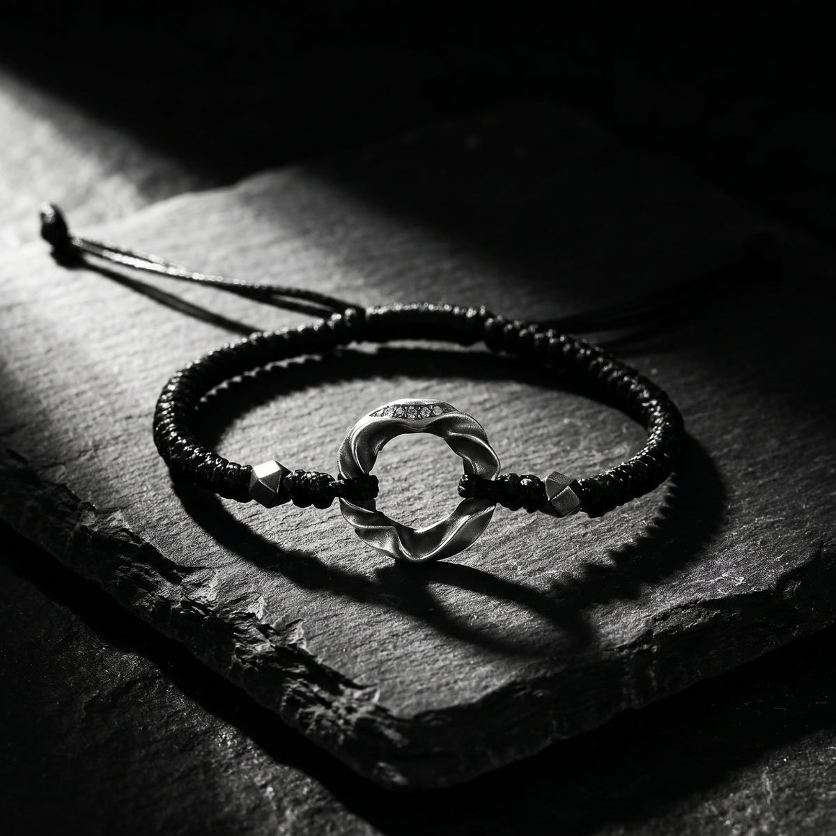 Black braided cord bracelet with a twisted silver ring and geometric beads, displayed flat on a textured slate slab.