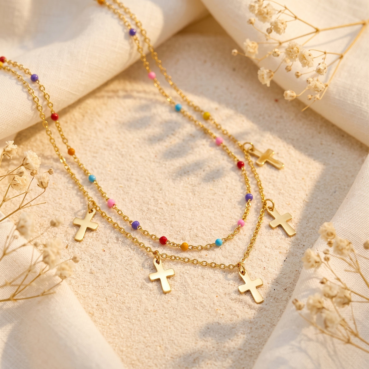 Close-up flat lay of the double-layer gold-tone necklace with colorful beads and dangling cross charms on sand.