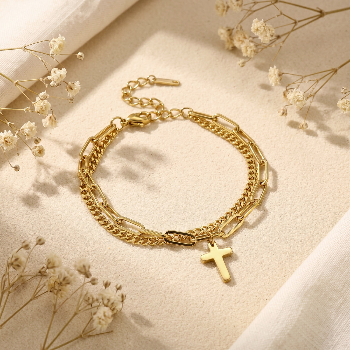 Gold-tone double-layer cross bracelet displayed flat on a beige surface with dried white flowers.