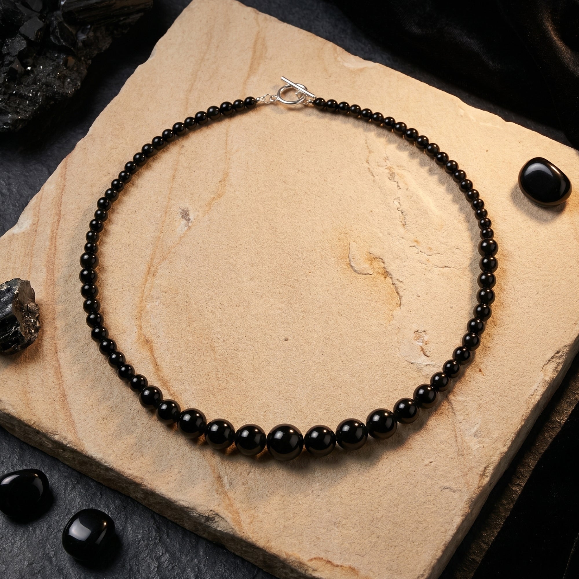 Top-down view of a graduated natural black onyx bead necklace with a silver toggle clasp on a textured stone slab.