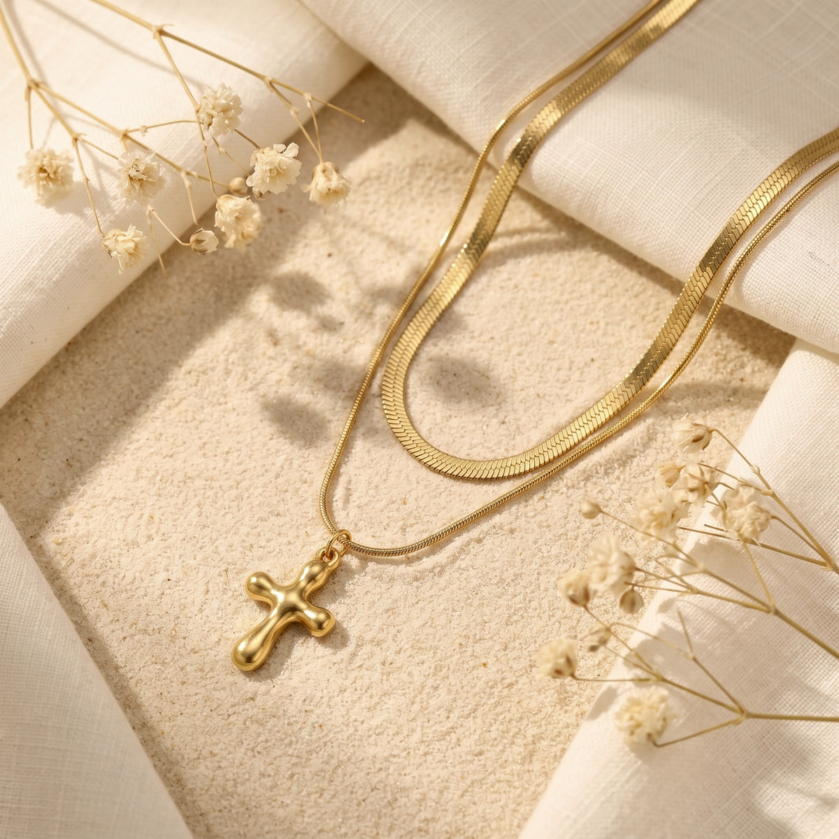 Gold-tone double-strand necklace resting on sand, with a flat chain, fine chain, and smooth cross pendant.