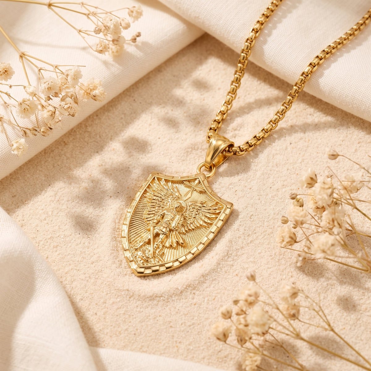 Gold-tone shield pendant necklace with sculpted archangel relief resting on sand beside dried flowers and white fabric.