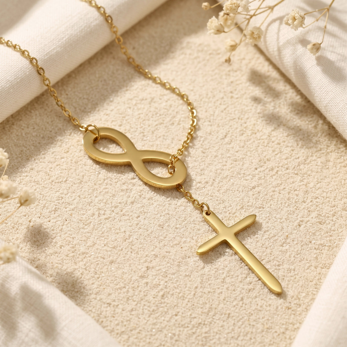 Gold-tone lariat necklace with an infinity pendant and hanging cross displayed on a textured beige surface