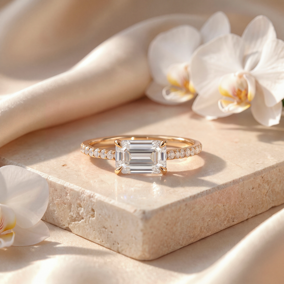 Yellow gold engagement ring with an east-west emerald-cut moissanite and pavé band on a beige stone surface.