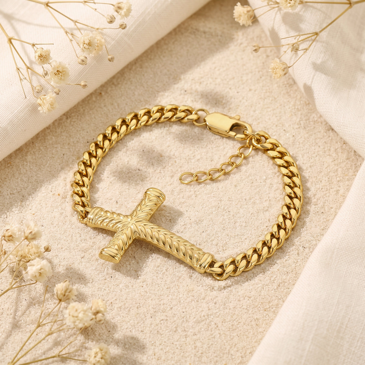 Gold-tone Cuban-link bracelet with a chevron-textured cross, clasp, and extender on a sandy surface.