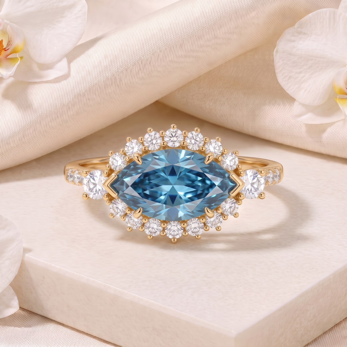 Marquise-cut blue moissanite halo ring in yellow gold set on a marble pedestal with silk and orchids.