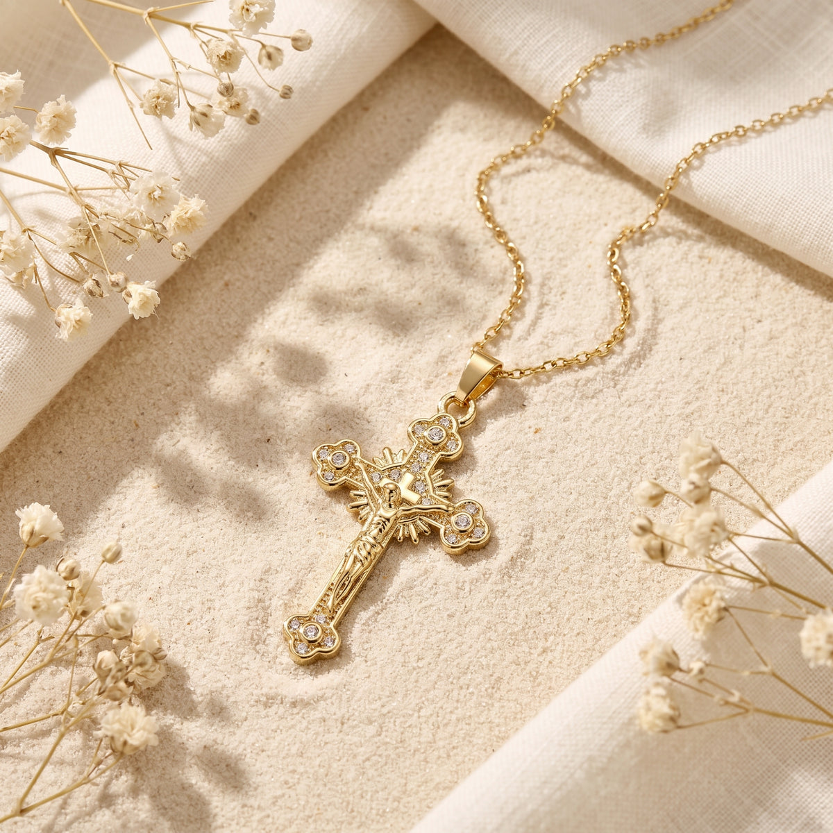 Gold-tone ornate crucifix necklace with clear crystal accents laid flat on sand with linen and dried flowers.