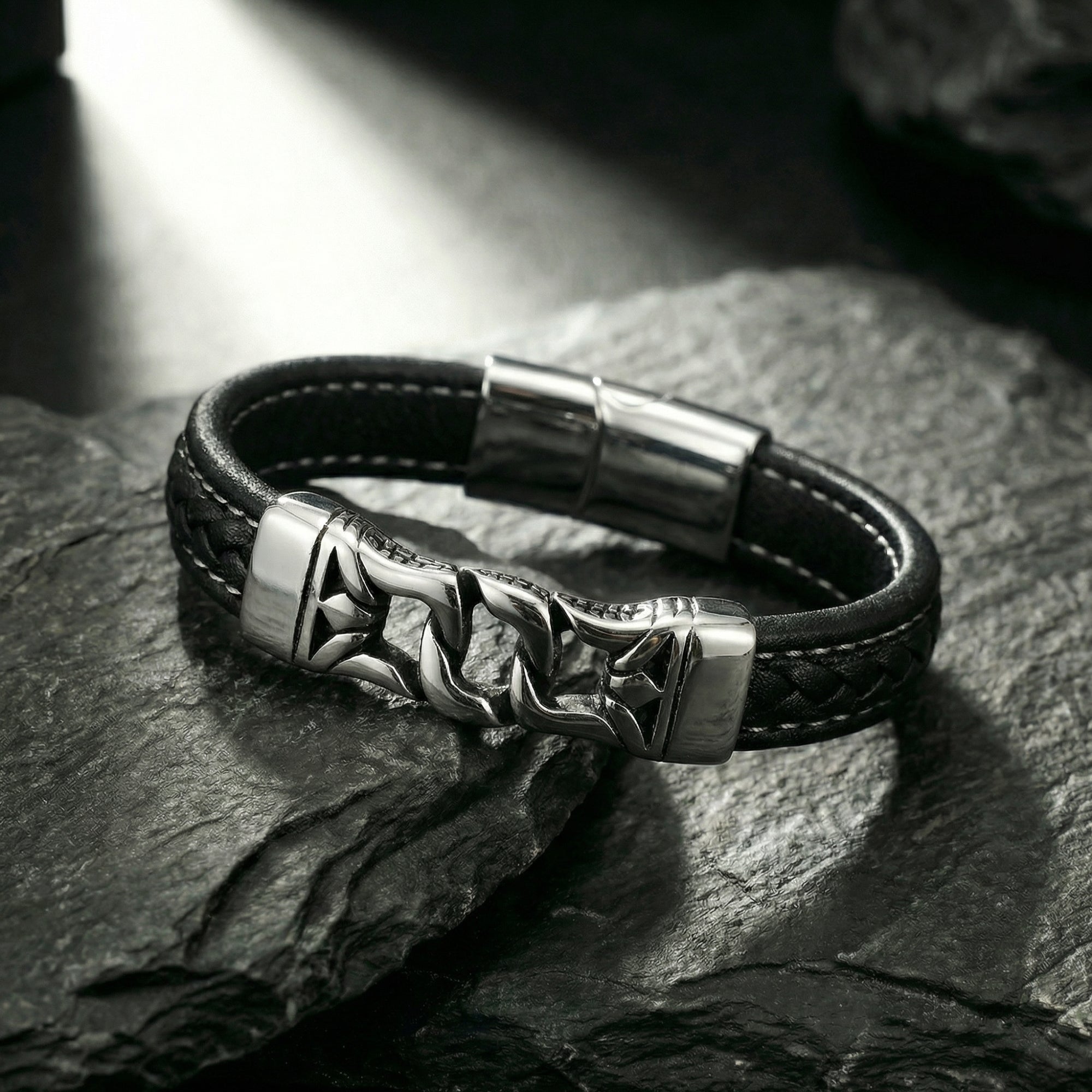 Black braided leather Nexus Bracelet with silver stainless steel centerpiece on a dark textured stone background.
