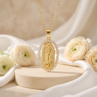 Gold-tone Guadalupe oval pendant necklace with cubic zirconia halo hanging above a pedestal and white roses.