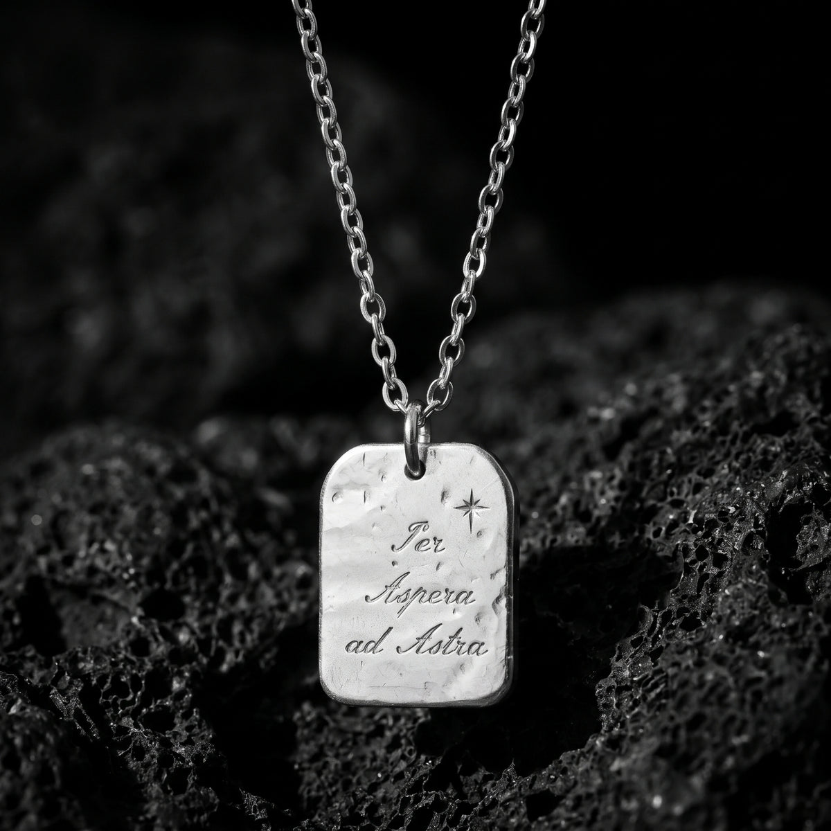 Silver tag pendant engraved with “Per Aspera ad Astra” and an eight-point star, suspended over dark porous rock.