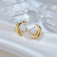 Gold-tone crescent moon stud earrings with round opal stones displayed on white silk with clear crystals.
