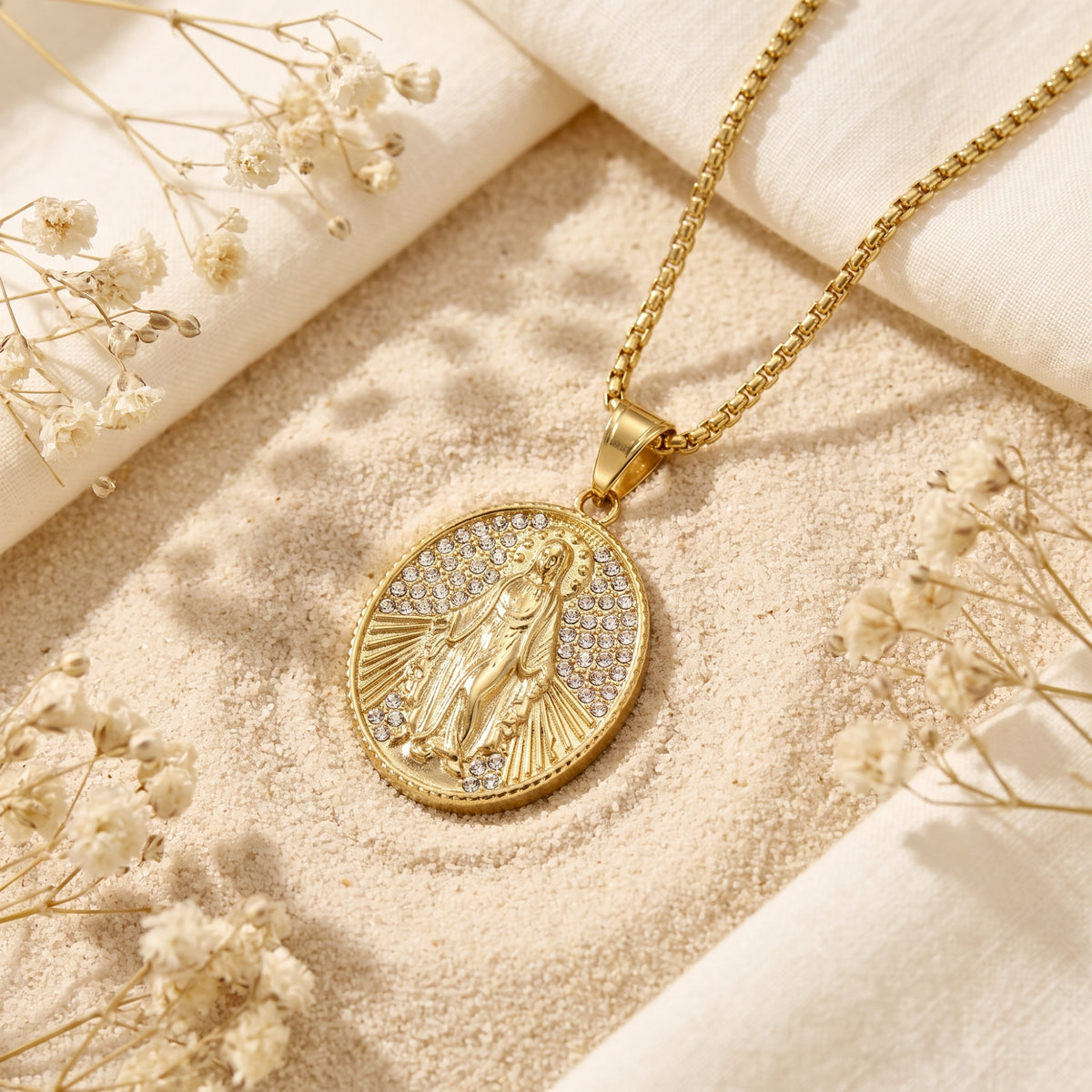 Close-up of a gold-tone oval Miraculous Medal necklace with clear stone accents resting on sand among dried flowers.