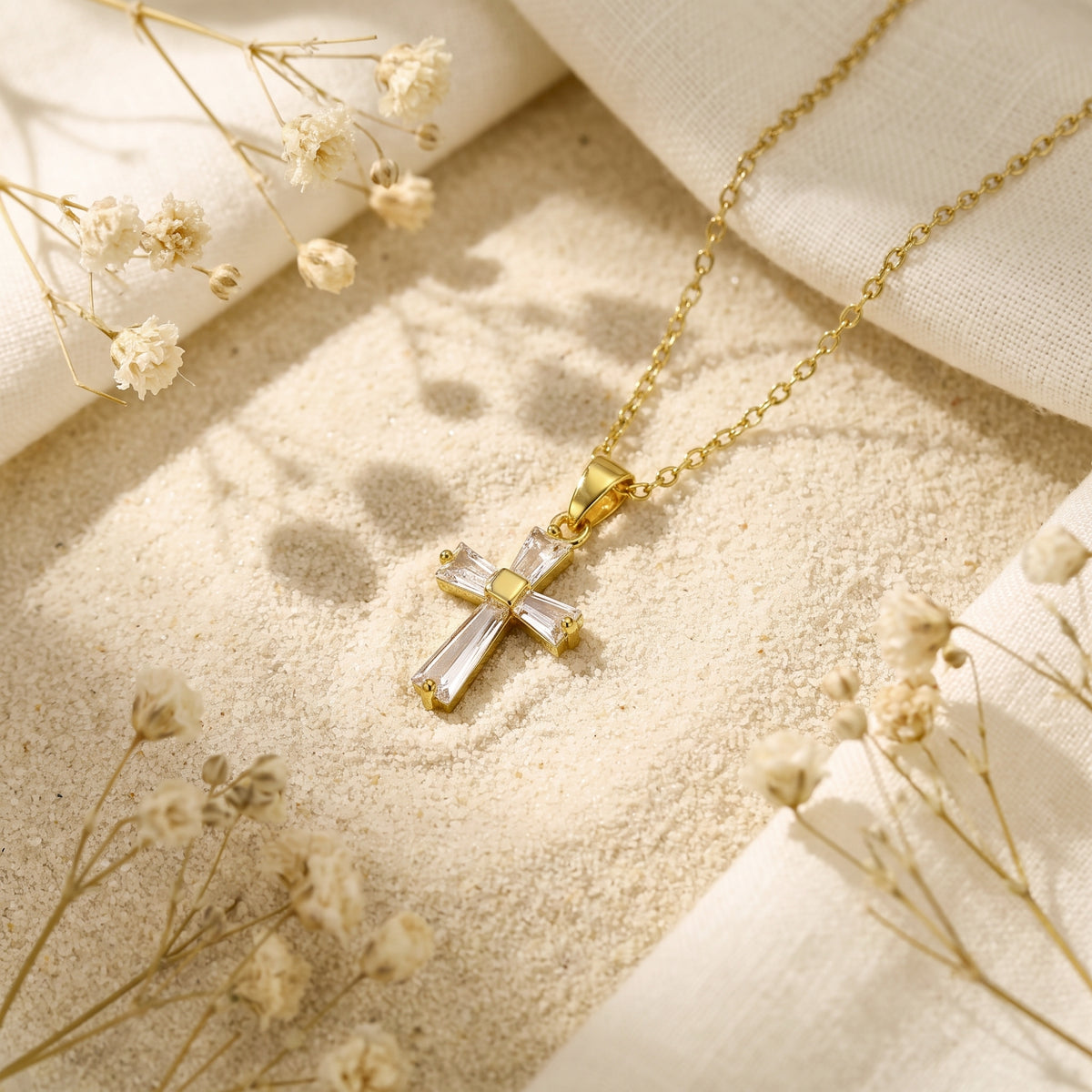 Gold cross necklace with clear baguette cubic zirconia, laid on sand beside white fabric and dried flowers.