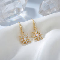 Gold-tone starburst drop earrings with round white opals on white silk with decorative crystals.