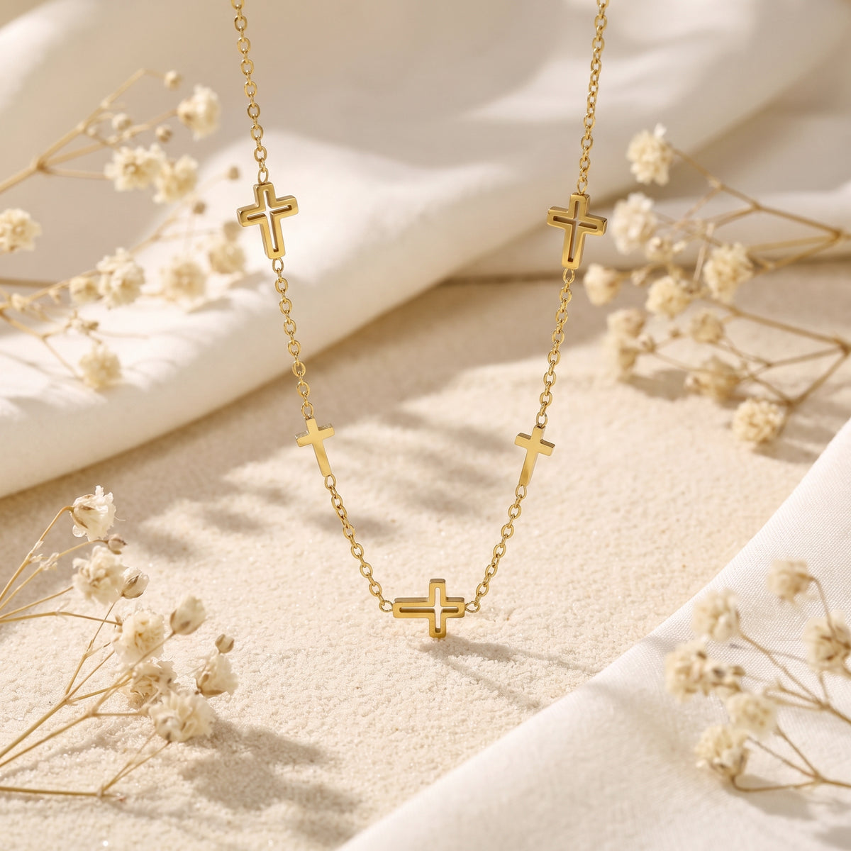Gold-tone multi-cross station necklace arranged in a U shape on textured sand, white fabric, and dried flowers.