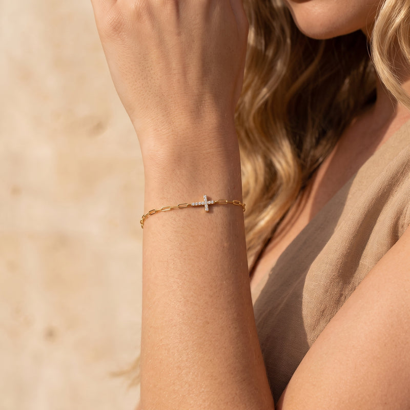 Gold-tone paperclip chain bracelet with a pave-set cross worn on a raised wrist.