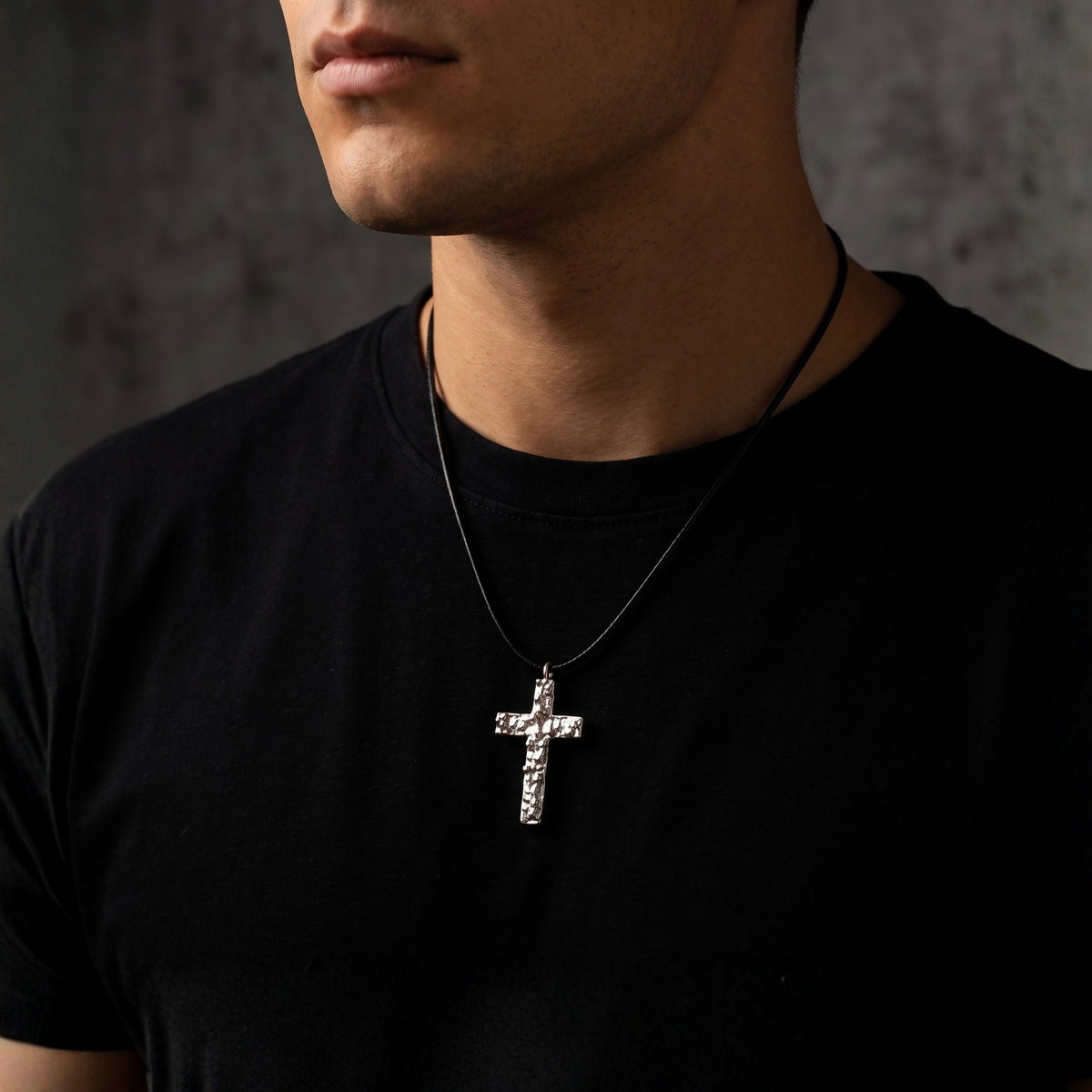 Model wearing a textured silver cross pendant on a black cord over a black T-shirt.