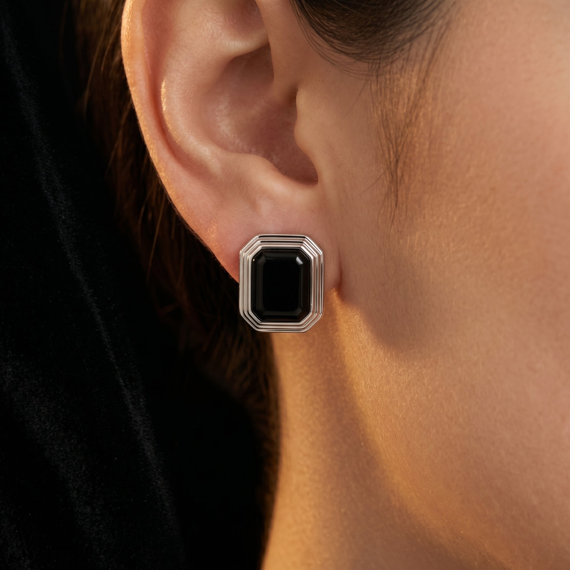 A square cut natural black onyx earring in silver worn on a woman's ear, showing its large scale and mirror-like finish.