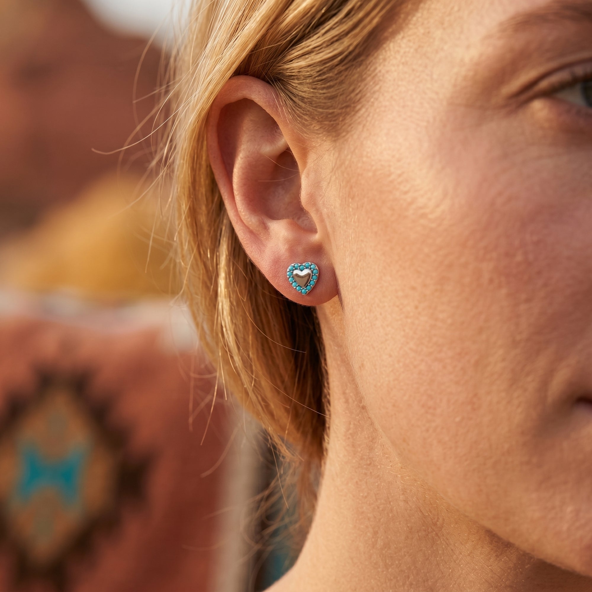 Close-up of a woman wearing a silver heart stud earring with turquoise stones in a Southwestern outdoor setting.