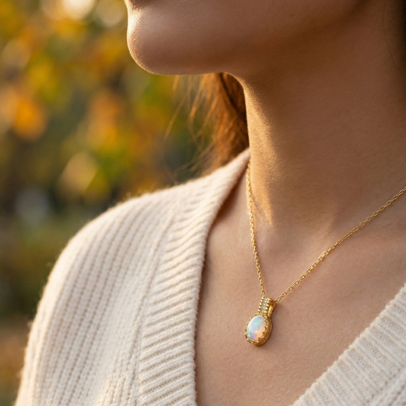 Model wearing the gold-tone Orianthi necklace with an oval white opal pendant against a blurred autumn background.