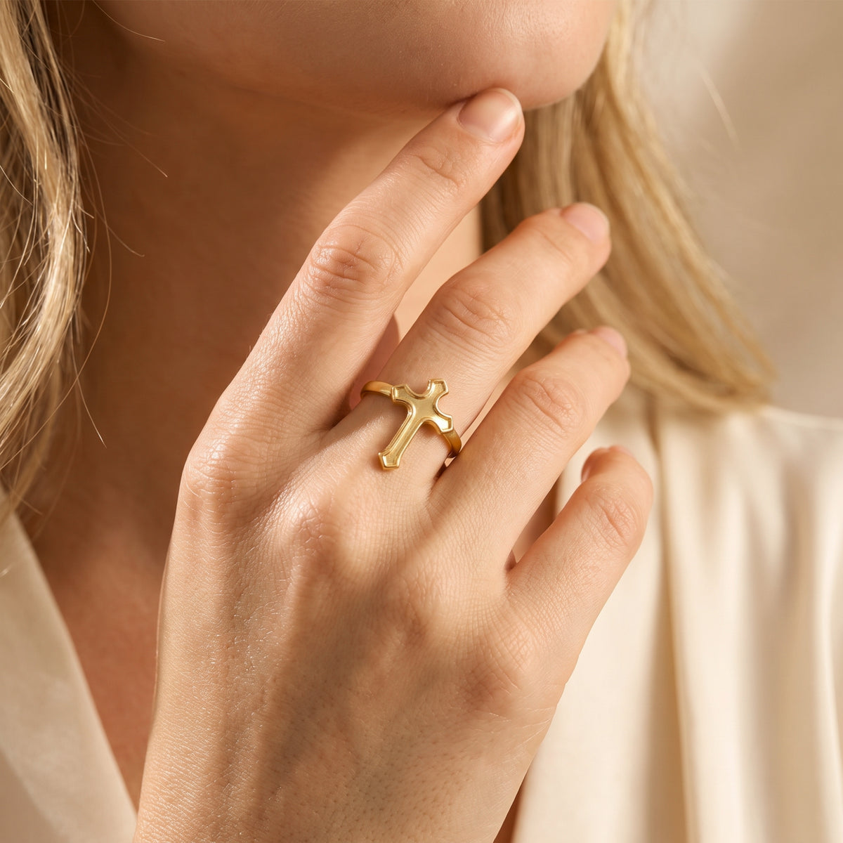 Gold-tone cross ring worn on a woman's finger beside her chin in a close-up lifestyle view.