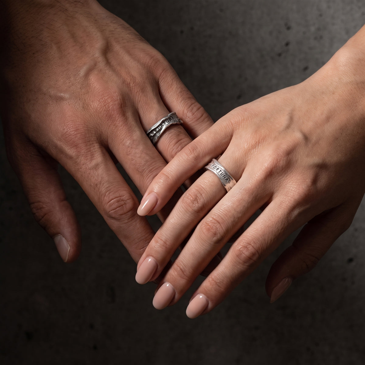 Two hands wearing textured open silver rings with FORTUNA inscriptions, one darker and one lighter.