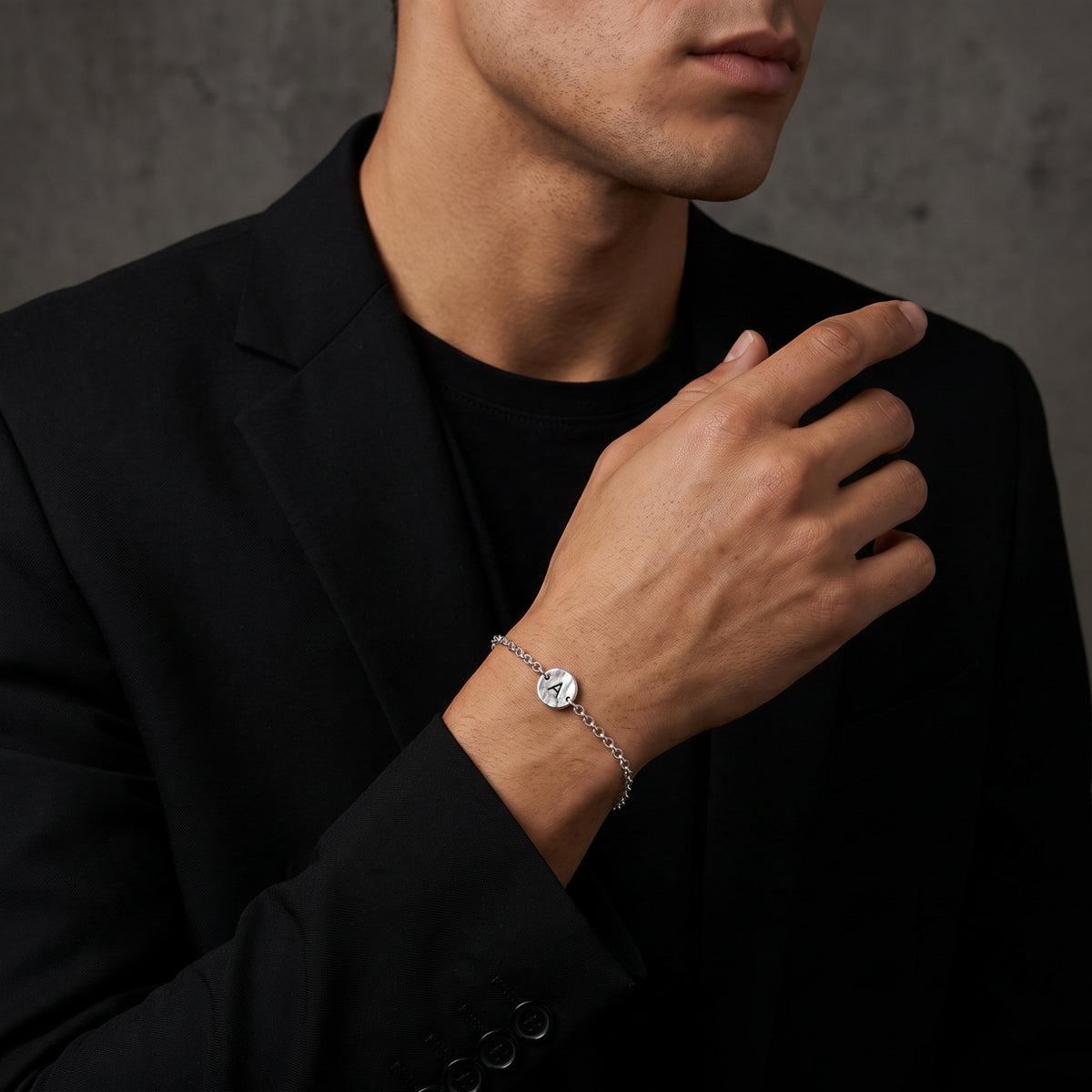Silver chain initial A bracelet worn on a man's wrist with a black suit jacket and crewneck shirt.