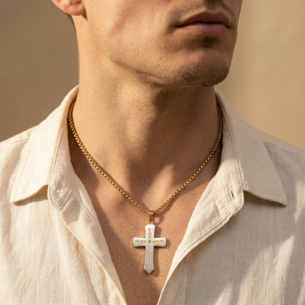 Man wearing a two-tone cross pendant on a gold-tone box chain with an open-collar cream shirt.