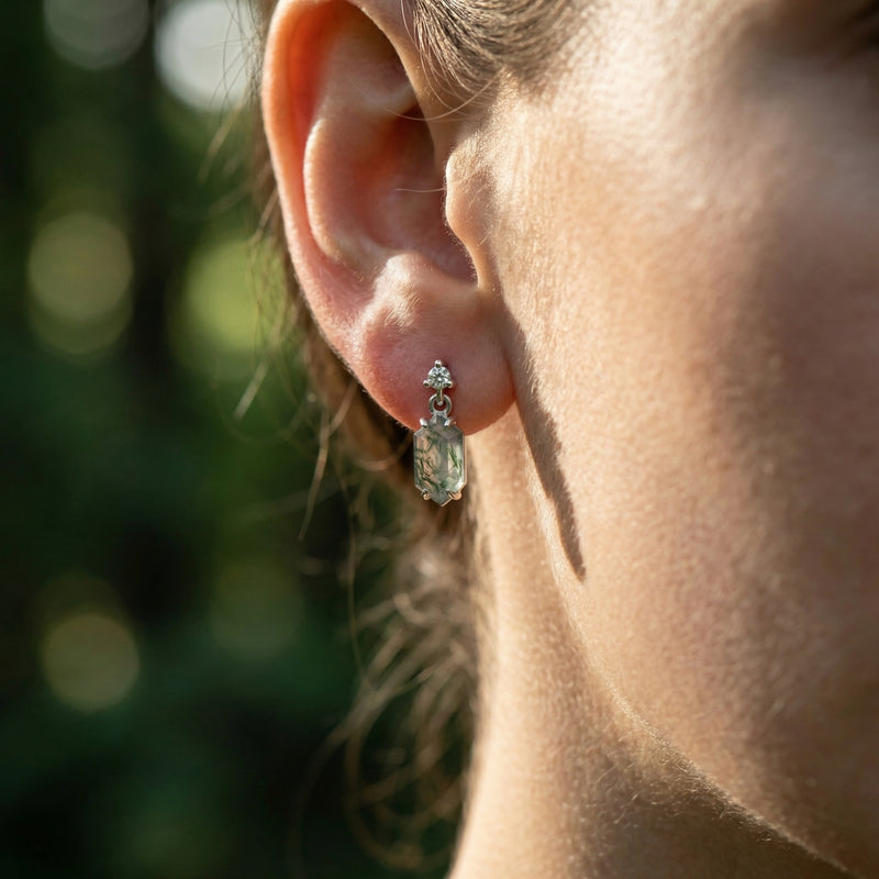 Side view of a barrel cut moss agate dangle earring in sterling silver worn on a model's ear.