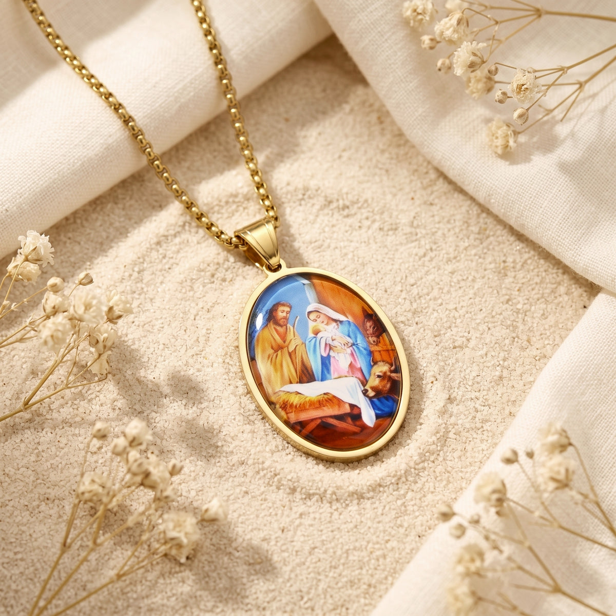 Gold-tone oval Nativity pendant necklace lying diagonally on pale sand beside white fabric and dried flowers.