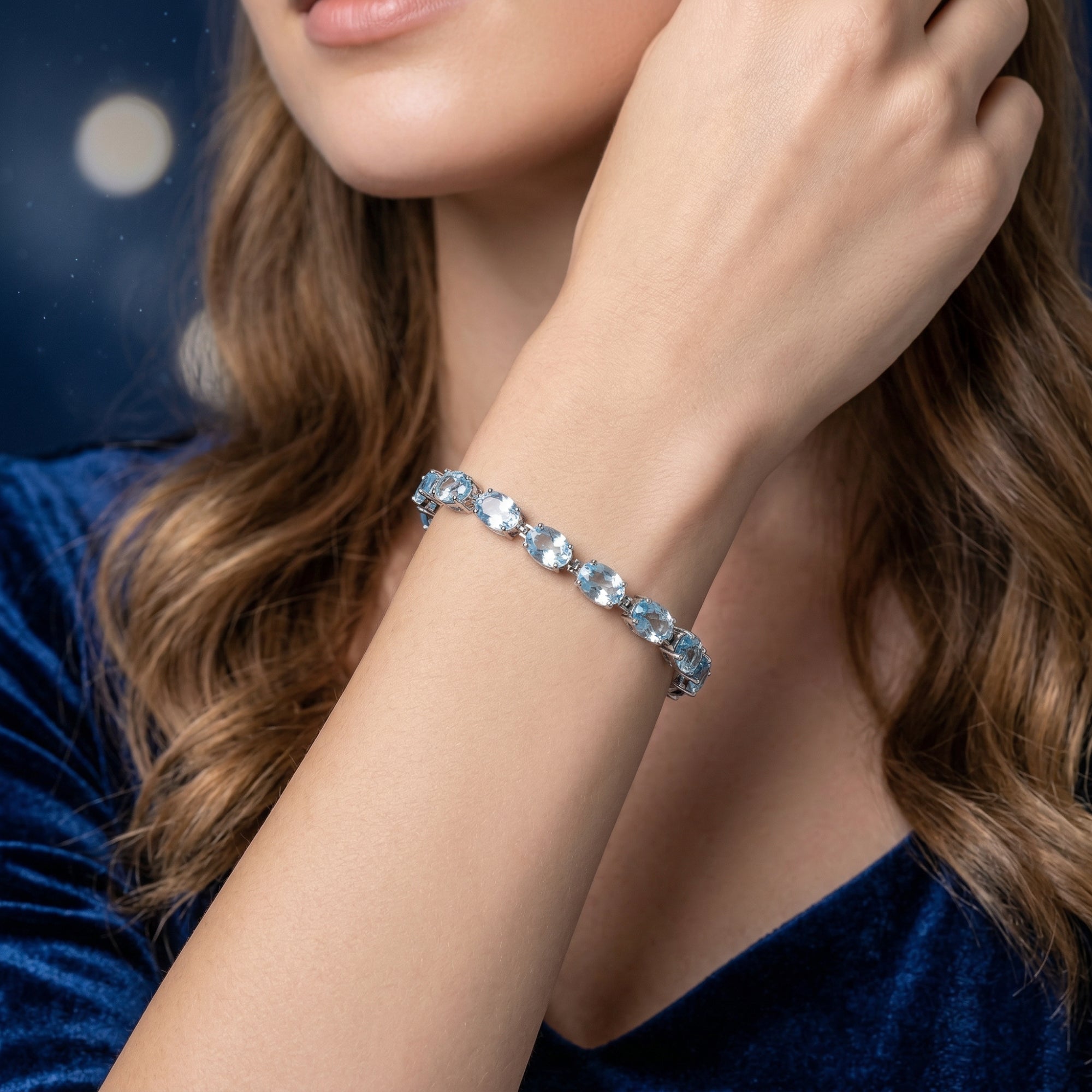 Woman wearing the oval cut blue topaz tennis bracelet in white gold on her wrist, styled with a blue dress.