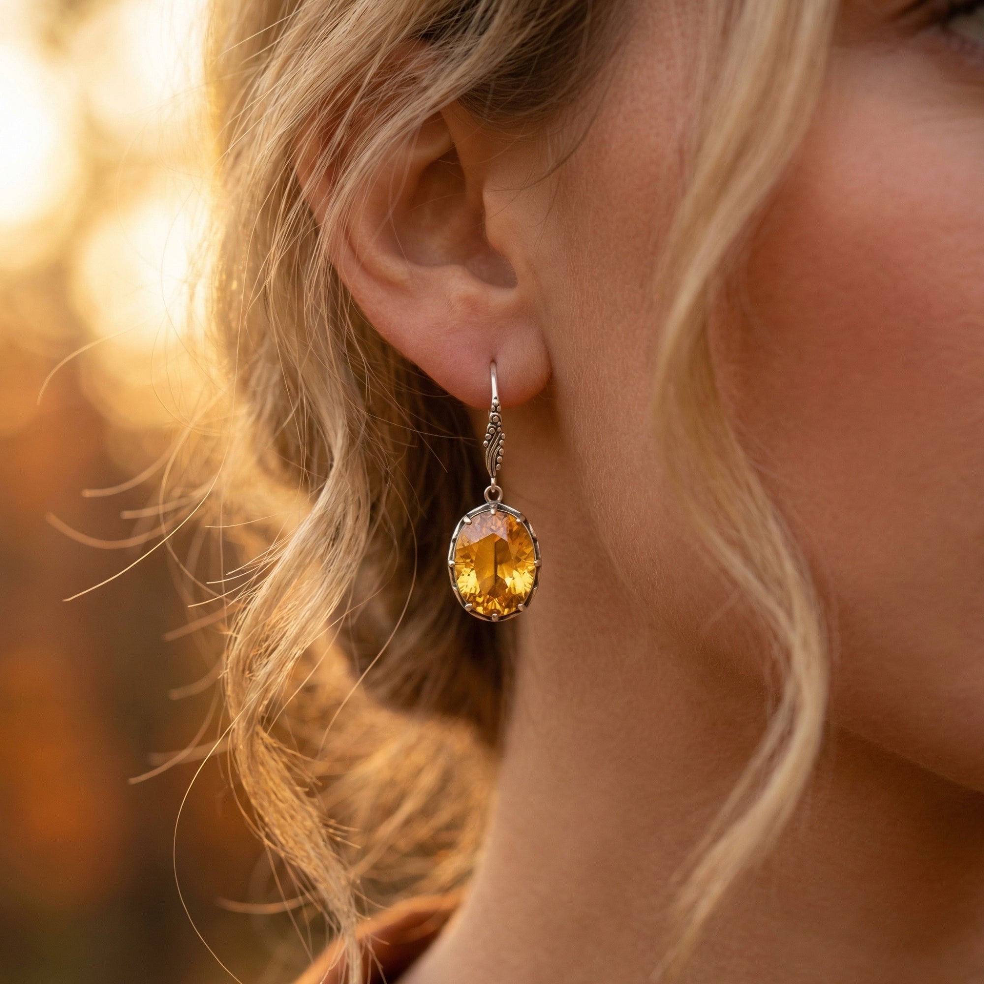Close-up of a silver oval cut citrine drop earring worn on a woman's ear during golden hour.