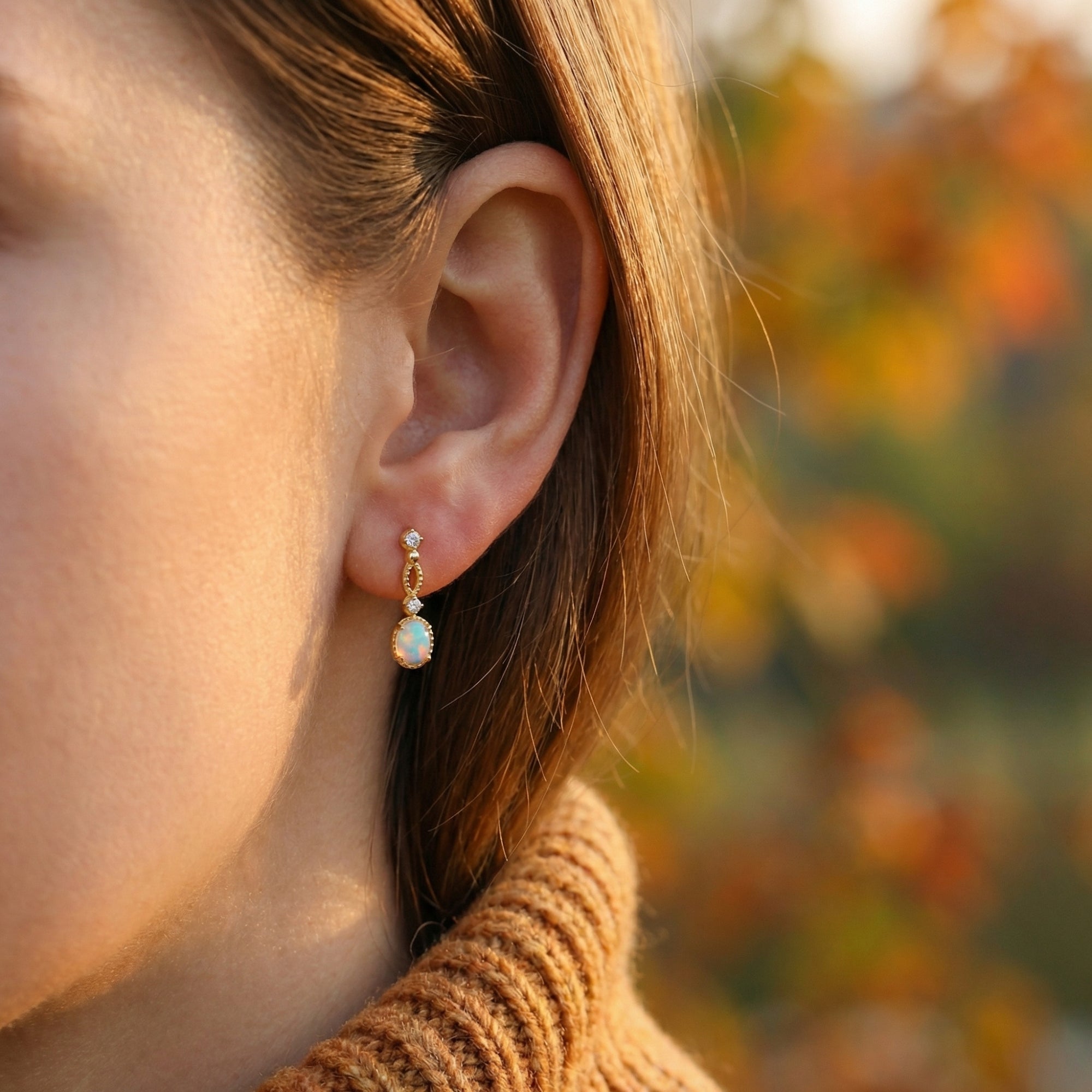 A woman wearing the gold-tone Lorelei oval white opal drop earring against a blurred autumn background.