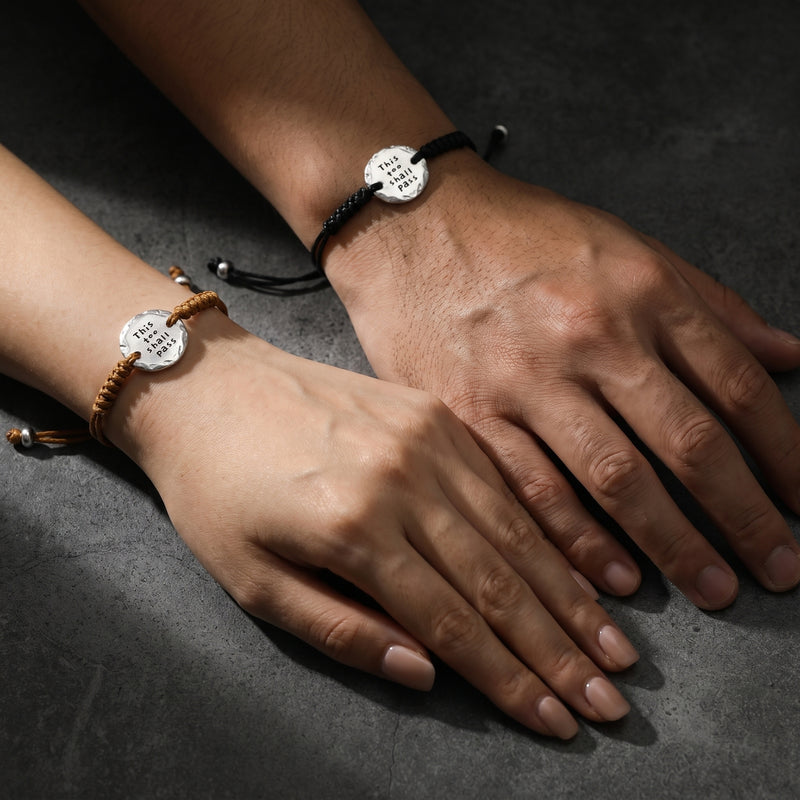 Two wrists wearing brown and black cord bracelets with silver medallions over a dark surface.