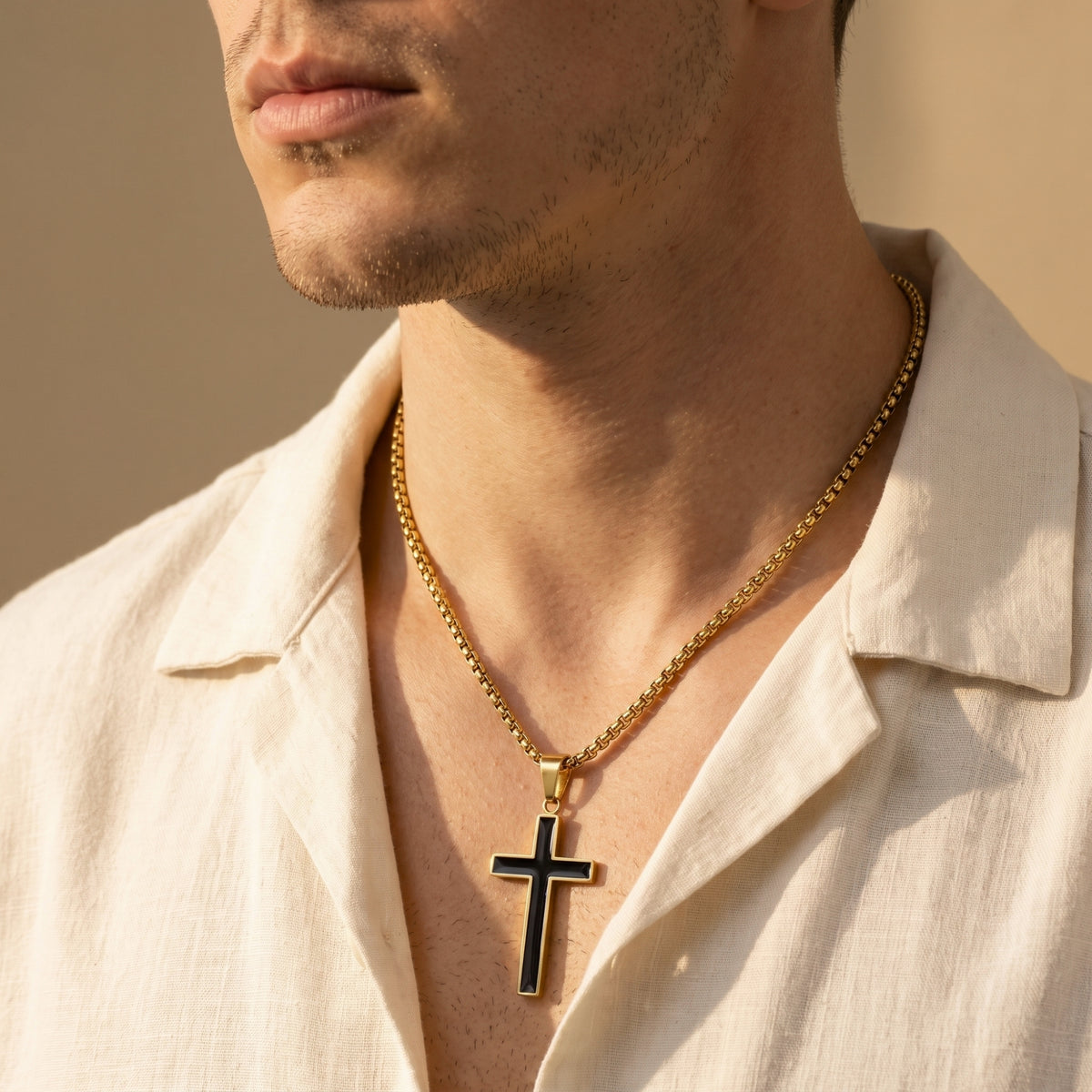 A man wears the gold-tone cross necklace with black enamel inlay and box chain over a cream shirt.