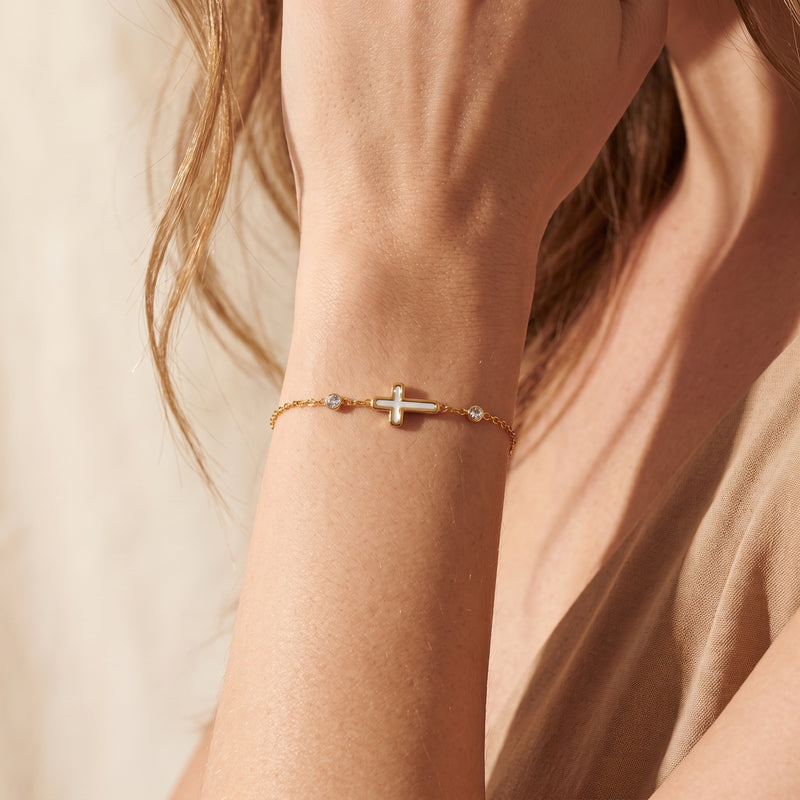 Close-up of the gold-tone station bracelet with a white cross worn around a person’s wrist.