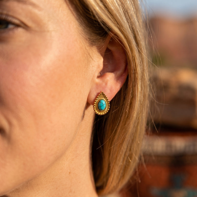 A woman wearing the Sundance turquoise drop earring with an 18K gold beaded border against a desert background.