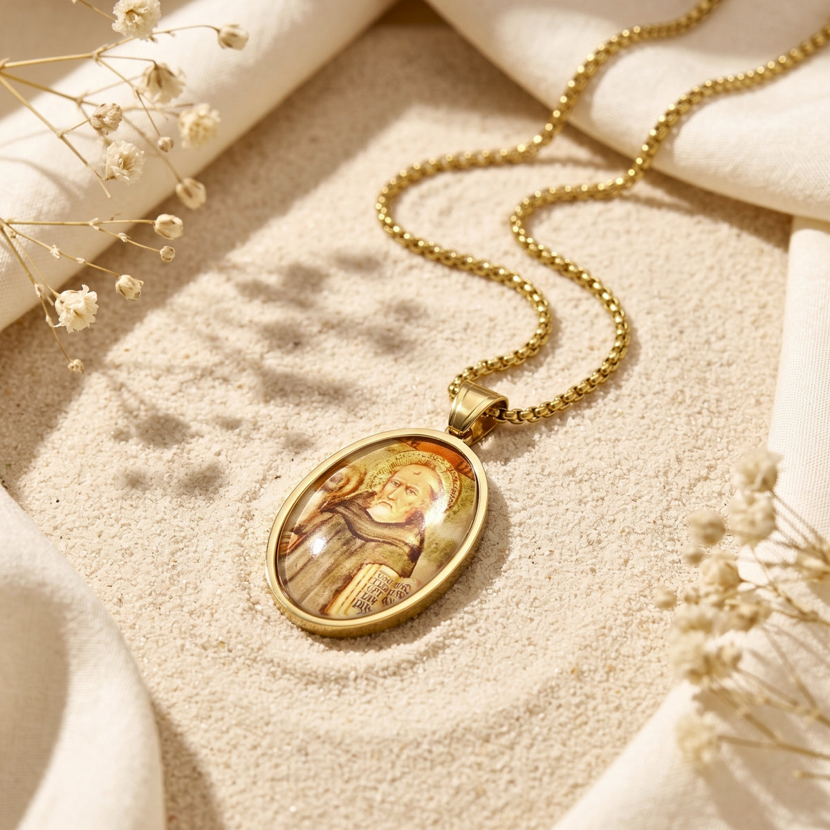 Gold-tone oval saint icon pendant necklace resting on light sand beside draped white fabric and dried flowers.
