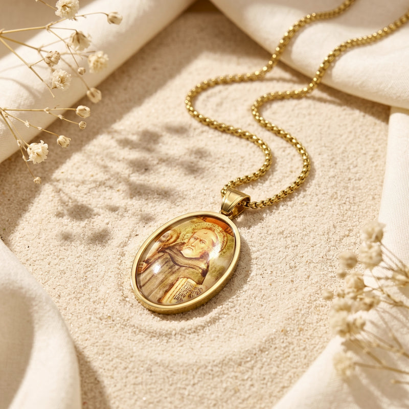 Gold-tone oval saint icon pendant necklace resting on light sand beside draped white fabric and dried flowers.