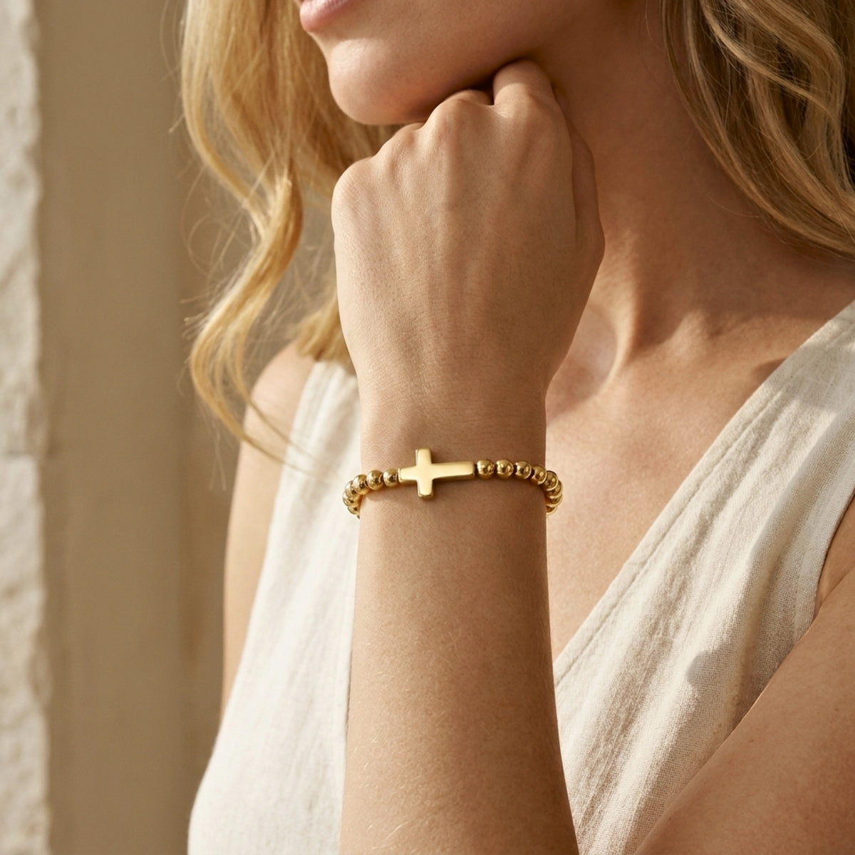 Model wearing a gold-tone beaded cross bracelet on her wrist with her hand resting beneath her chin.