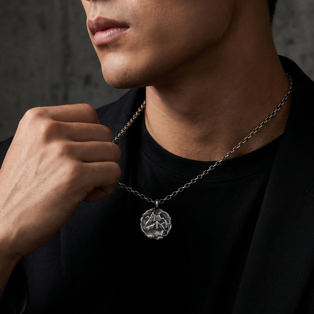 Close-up side view of a model holding a silver mountain-relief pendant over a black shirt and jacket.