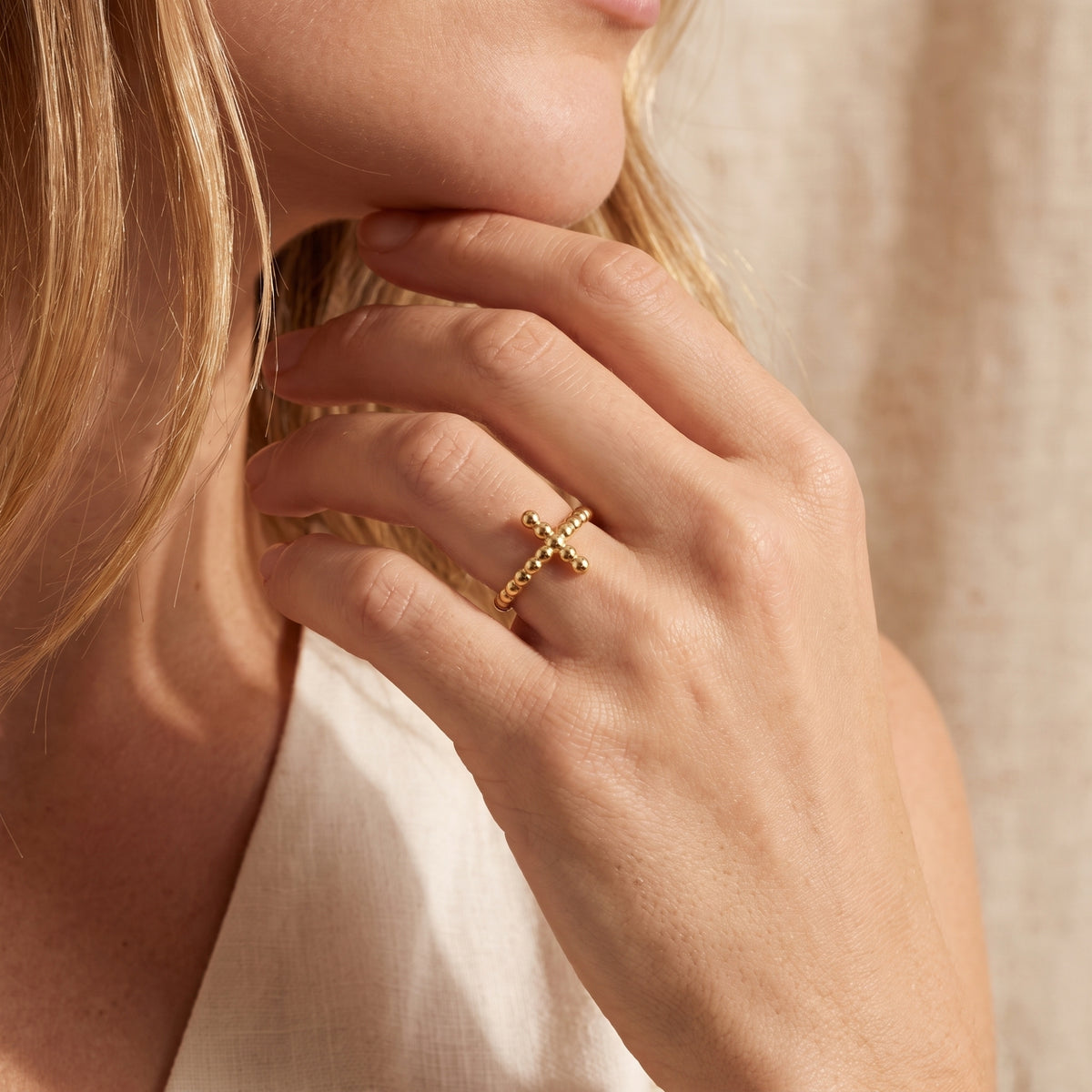 Close-up of a gold-toned beaded cross ring worn on a finger, with the hand resting near the model’s chin.