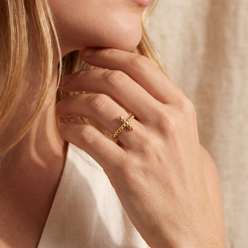 Close-up of a gold-toned beaded cross ring worn on a finger, with the hand resting near the model’s chin.