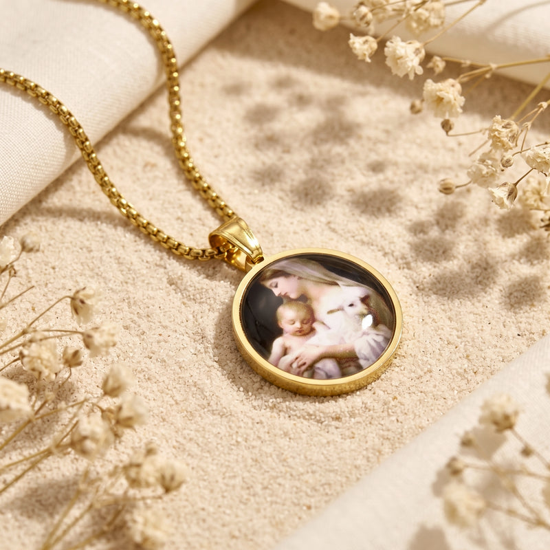 Angled view of the gold-tone Madonna and Child pendant resting flat on fine sand alongside dried flowers.