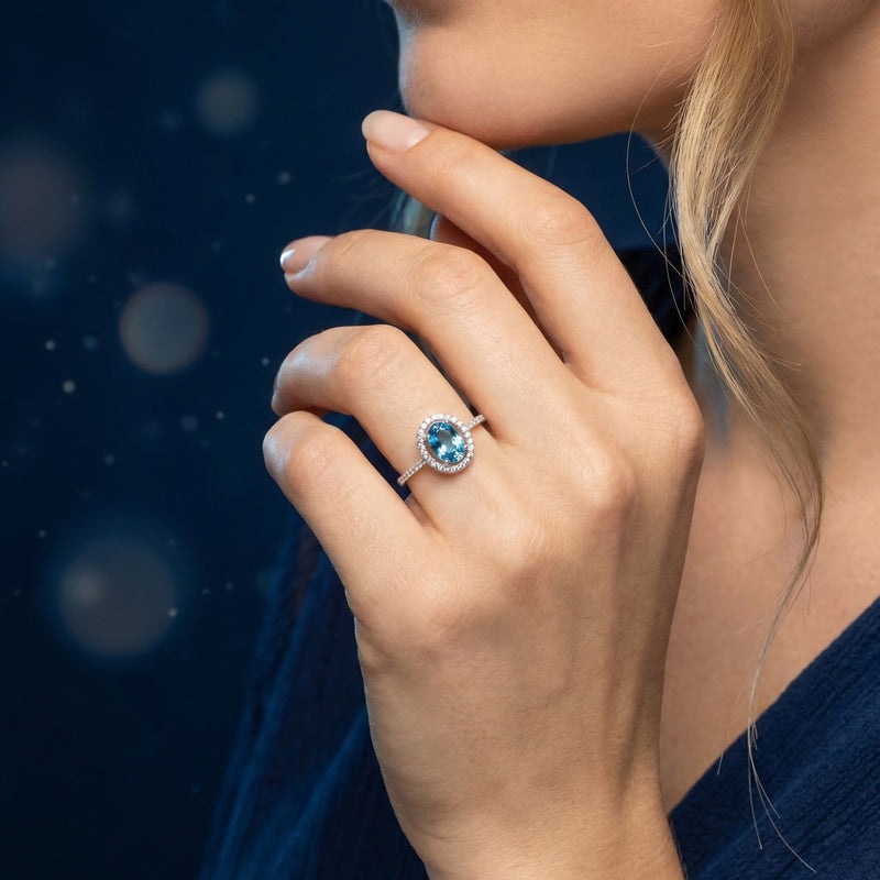 Sterling silver oval blue topaz halo ring worn on a model's hand against a dark blue bokeh background.