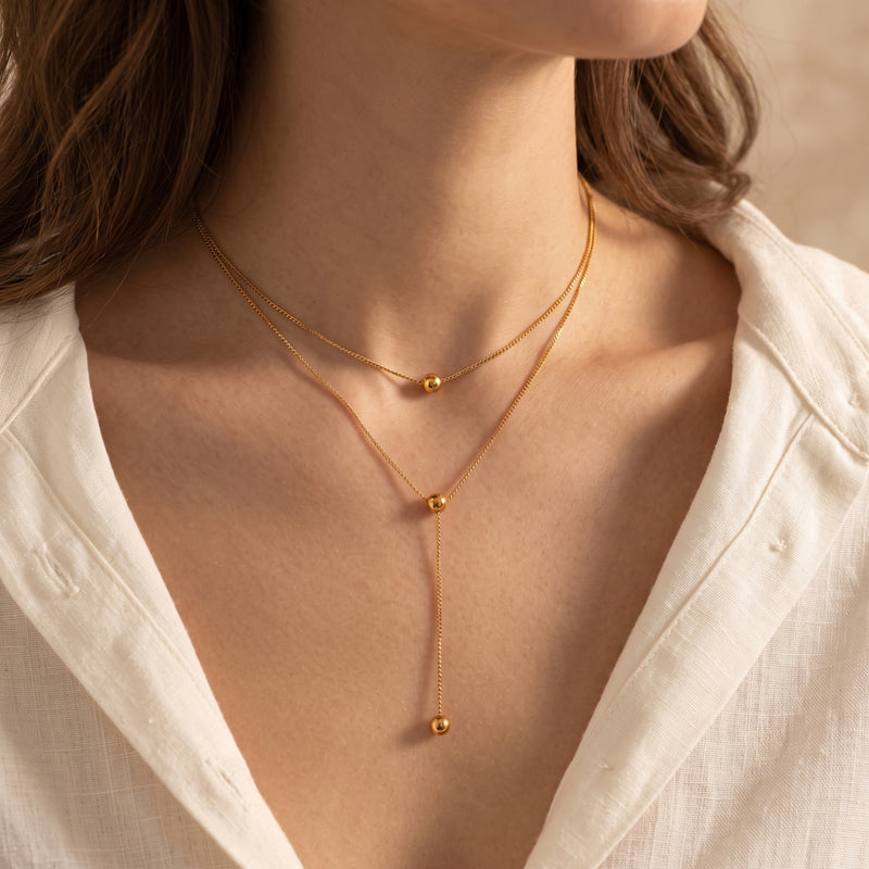 Gold layered lariat necklace with polished spheres worn by a woman in a white button-down shirt.