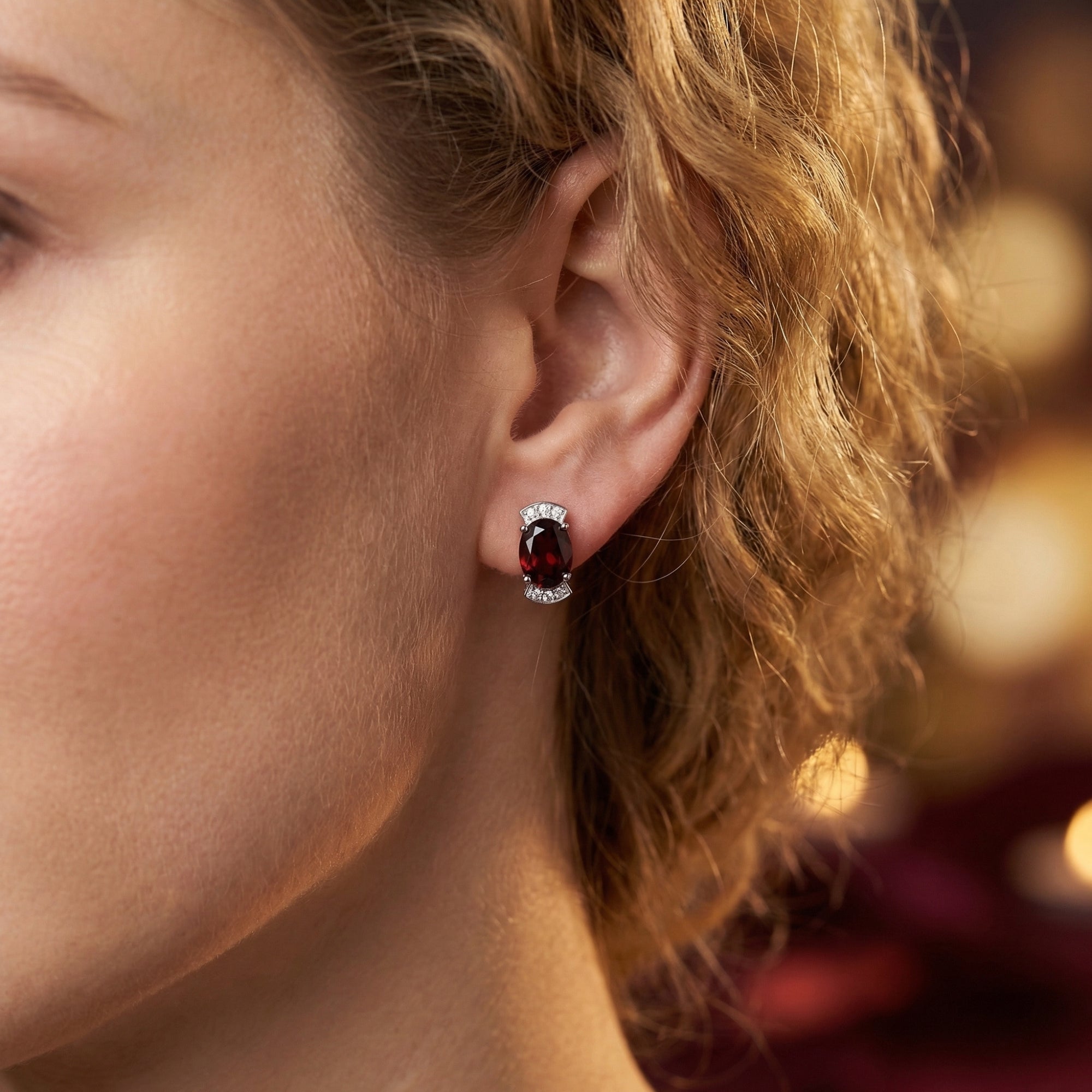 Woman wearing a Bellissima oval natural garnet drop earring in white gold against a warm bokeh background.
