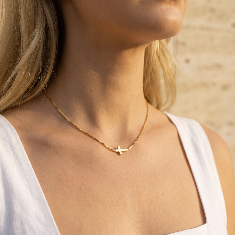 Model wearing a gold-tone cross necklace on a box chain above a white top, shown from the neck down.